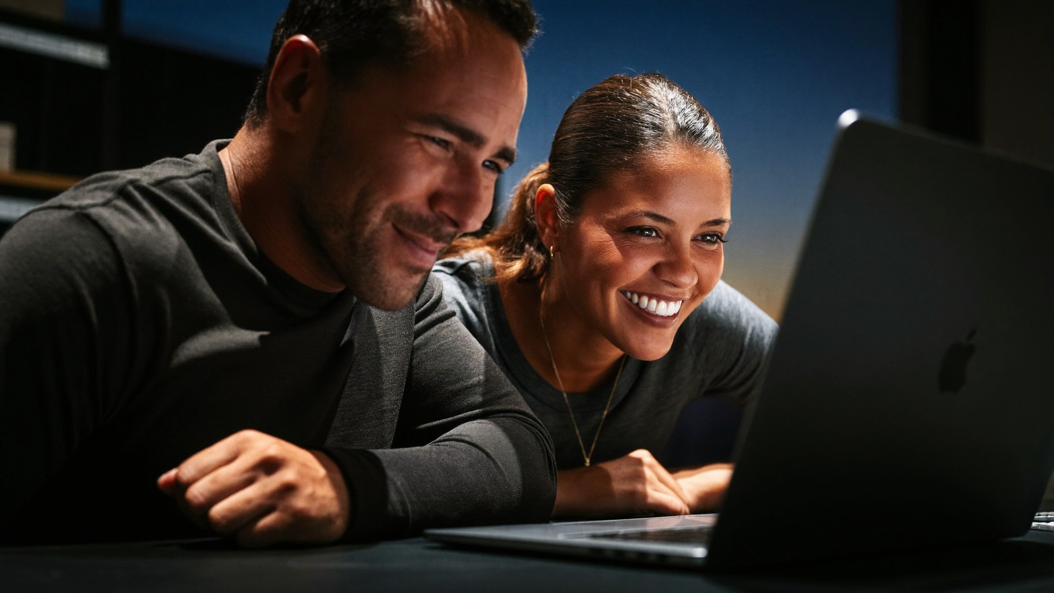Two people smiling at the same laptop screen, close and warmly lit.